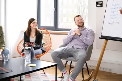 Workshop im Meetingraum Mitarbeiter:innen sitzen im Meetingraum, einer zeigt auf ein Flipchart