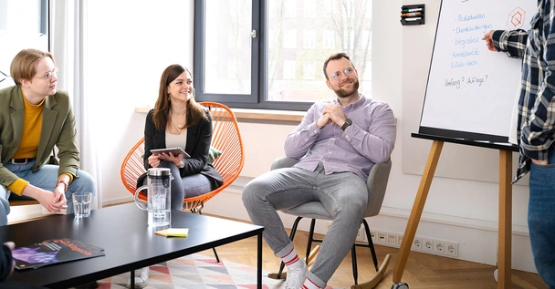 Workshop im Meetingraum Mitarbeiter:innen sitzen im Meetingraum, einer zeigt auf ein Flipchart