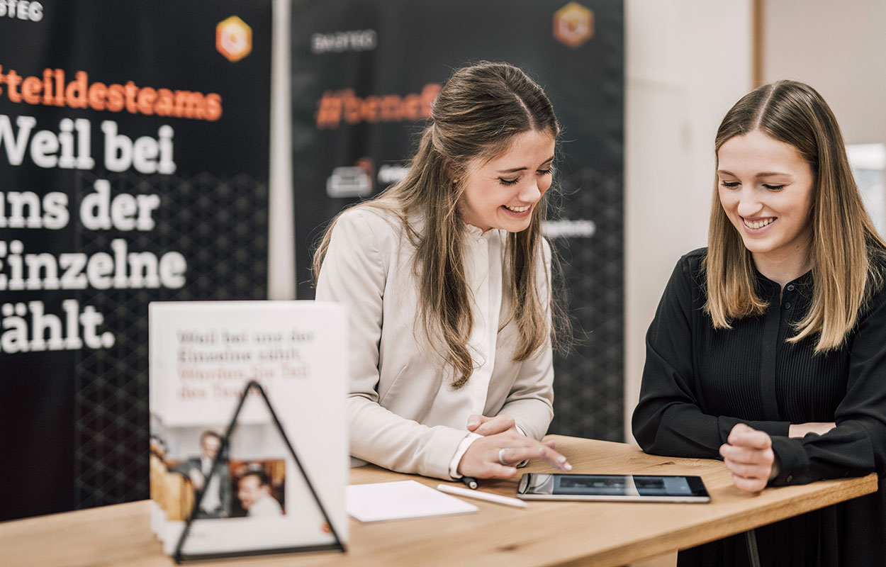 Zwei HR-Mitarbeiterinnen stehen an einer Ecke des Tisches vom Babtec-Stand und betrachten lächelnd gemeinsam den Screen eines Tablets.